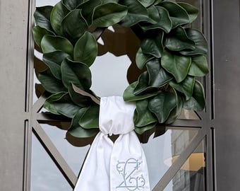 Puede incluir: Una exuberante corona de hojas de magnolia verde con una cinta blanca con un monograma bordado. La corona se exhibe en una puerta de metal oscuro con un diseño decorativo, creando una estética elegante y acogedora.