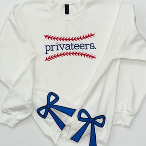 以下が含まれることがあります： 赤い野球のステッチデザインに青い文字で「privateers」と書かれた白いスウェットシャツ。袖には青いリボンが付いています。