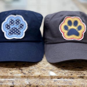 Op de afbeelding: Vier baseballpetten in lavendel, marineblauw, houtskool en geel. Elke pet heeft een pootafdrukontwerp in verschillende kleuren. De pootafdrukken zijn geborduurd met een contrasterende omtrek.