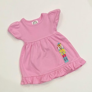 May include: A light pink dress with short, ruffled sleeves and a ruffled hem. The dress features an embroidered nutcracker design on the front. The dress has a round neckline and a gathered waist.