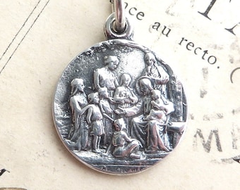 May include: Silver pendant featuring a relief sculpture of a group of children gathered around a seated figure. The pendant is circular and has a slightly textured surface.