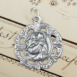 May include: Silver pendant featuring a detailed image of Saint Anthony of Padua holding the Christ Child. The pendant has a decorative, openwork design around the image.