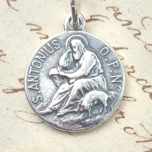 Può includere: Ciondolo argentato con un rilievo dettagliato di una figura seduta con un'aureola, un maiale e l'iscrizione "S. ANTONIUS O.P.N." Il ciondolo ha un piccolo anello nella parte superiore per una catena.