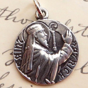 May include: Silver charm featuring a detailed image of Saint Francis of Assisi holding a bird in his hand. The charm is round and has a loop for attaching to a necklace or bracelet.