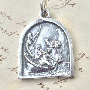 Op de afbeelding: Zilveren hangerbedel met een gedetailleerde reliëfsculptuur van drie figuren in een boot. De figuren zijn afgebeeld in een dynamische pose, wat beweging en actie suggereert. De hanger heeft een afgeronde, gebogen bovenkant en een gladde, gepolijste afwerking.