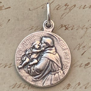 May include: Silver pendant featuring a relief of Saint Anthony of Padua holding a baby Jesus. The pendant has the inscription "S. ANT. PAD. ORA PRO NOBIS".