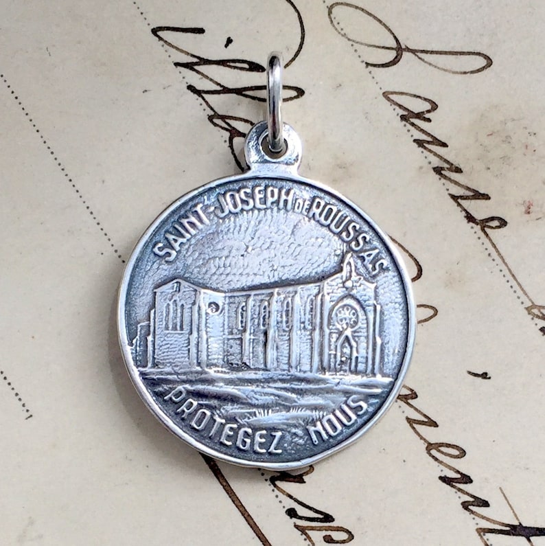 May include: Silver pendant charm featuring a detailed image of a church with the text "SAINT JOSEPH DE ROUSSAS PROTEGEZ NOUS".