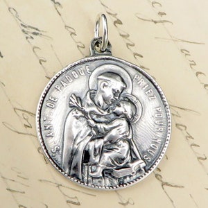 May include: Silver pendant featuring a relief image of Saint Anthony of Padua holding a baby. The pendant is inscribed with the text "Saint de Padoue, Priez pour nous".