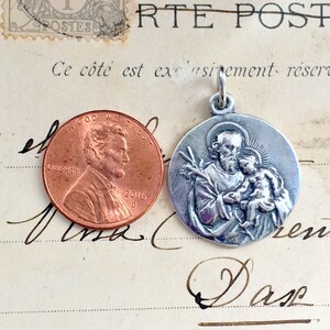 May include: Silver pendant featuring a detailed depiction of Saint Anthony of Padua holding the Christ Child. The pendant is shown next to a US penny for size comparison.