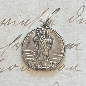 May include: A silver pendant featuring a detailed relief of a saint holding a staff. The pendant is round with the words "SAINT PATRICK" inscribed around the edge. The pendant has a small loop at the top for a chain.