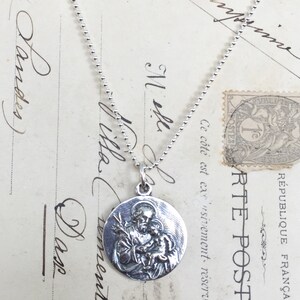 May include: Silver necklace with a round pendant featuring a detailed image of Saint Anthony holding a baby Jesus. The pendant is on a silver chain.