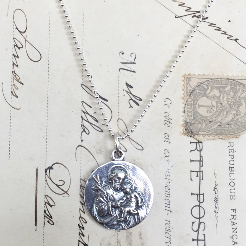 May include: Silver necklace with a round pendant featuring a detailed image of Saint Anthony holding a baby Jesus. The pendant is on a silver chain.