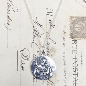 May include: Silver necklace with a round pendant featuring a detailed image of Saint Anthony of Padua holding a baby Jesus.