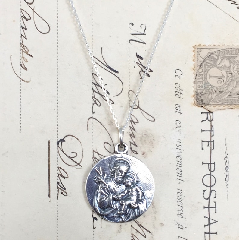 May include: Silver necklace with a round pendant featuring a detailed image of Saint Anthony of Padua holding a baby Jesus.