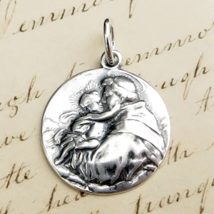May include: Silver pendant featuring a detailed relief sculpture of a saint holding a child. The pendant is round and has a loop at the top for attaching to a chain.