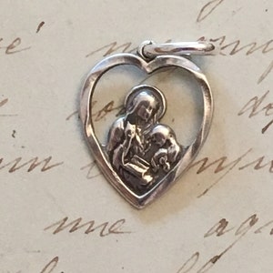 May include: A silver heart-shaped pendant with a religious motif. The pendant shows a figure holding a child within the heart's outline. It has a small loop at the top for attaching to a chain.