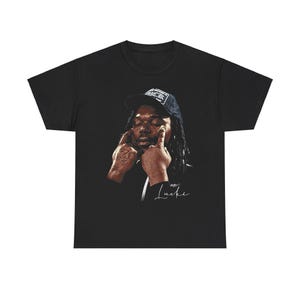May include: Black t-shirt with a graphic of a man wearing a black baseball cap and a white signature that says "w/c/ Luckie".