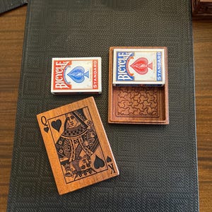 May include: Two wooden boxes with a playing card design. The boxes are open and show a red and blue Bicycle playing card inside. The boxes are on a black surface with a woven pattern. The third box is closed and shows a Queen of Spades playing card design.