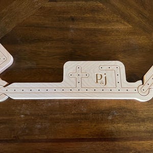 Pode incluir: Um tabuleiro de jogo de madeira clara com as iniciais "Pj" gravadas. O tabuleiro tem uma forma única com várias seções conectadas por dobradiças e linhas pontilhadas. O tabuleiro está em uma superfície de madeira escura.