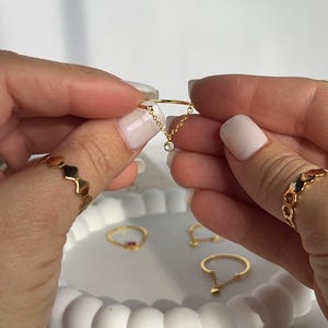 Op de afbeelding: Een delicate gouden ring met een klein kettinkje en een enkele heldere steen die eraan hangt. De ring wordt getoond aan een vinger en op een witte displaystandaard met andere ringen.