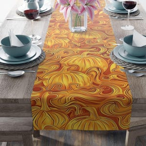 May include: A decorative table runner featuring a repeating pattern of stylized pumpkins in shades of orange and yellow. The runner is placed on a wooden table, with place settings and a vase of pink flowers visible.