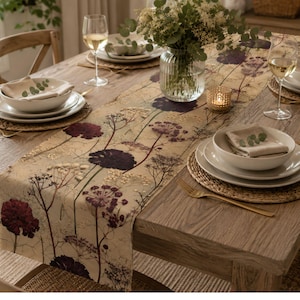 May include: A dining table set with a floral table runner featuring burgundy and cream botanical patterns. White plates, bowls, and gold cutlery are set on woven placemats. A glass vase holds greenery, and wine glasses are filled with a light-coloured beverage.