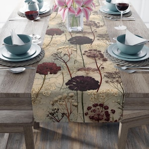 May include: A beige table runner with a floral pattern featuring dried flowers in shades of burgundy, purple, and white. The runner is laid out on a wooden table with place settings on either side.