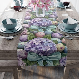 May include: A pastel purple and green table runner with a floral design featuring hydrangeas and speckled Easter eggs. The runner is decorated with a blue ribbon bow.