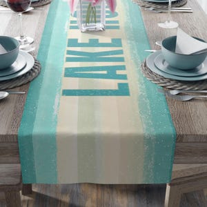 May include: A rectangular table runner with a striped design in shades of teal, cream, and white. The word "LAKE" is printed vertically in large teal letters. The runner is set on a wooden table with place settings.