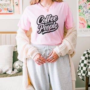 Peut inclure: Un t-shirt rose avec le texte "Coffee Before People" en lettres d'écriture noires.