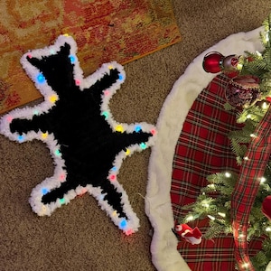 May include: A black bear-shaped rug with a white border and colorful lights. The rug is on a brown carpet next to a Christmas tree with red and white plaid tree skirt and ornaments.