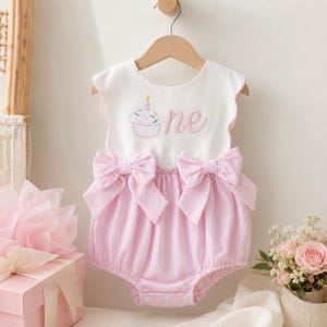 May include: A white sleeveless romper with a scalloped neckline and an embroidered cupcake graphic featuring the word one. The bottom portion is made of pink and white pinstriped fabric with two large bows at the waist and snap closures at the bottom.