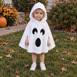 Op de afbeelding: Een witte poncho met capuchon voorzien van een zwarte spookgezicht-opdruk met twee ovale ogen en een ovale mond. Het kledingstuk heeft een puntige capuchon en zijsplitten voor armbeweging, gedragen over witte schoenen.