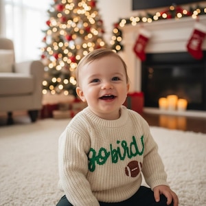 May include: A smiling baby wearing a cream-colored sweater with the words "Go Birds" in green script and a brown football graphic. The background includes a decorated Christmas tree, a fireplace with stockings, and a soft rug.