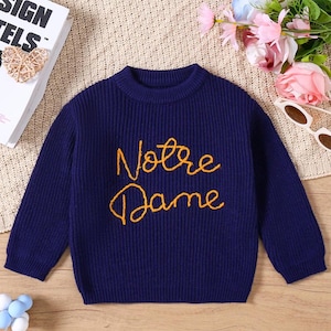 Op de afbeelding: Een marineblauwe trui met lange mouwen en een ronde hals. De trui heeft de woorden "Notre Dame" in goudkleurig script over de borst geborduurd. De trui is gemaakt van een geribbelde gebreide stof.