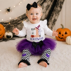 Könnte beinhalten: Ein weißer Langarmbody mit der Aufschrift My 1st Halloween, verziert mit Kürbis, Spinne und Hexenhut. Das Set enthält einen violetten Tüll-Tutu mit schwarzer Schleife sowie eine schwarz-weiß gezackte Leggings mit aufgenähten violetten Herz-Applikationen.