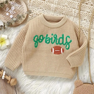 Könnte beinhalten: Beigefarbener Strickpullover mit dem grünen Schriftzug "go birds" und einer braunen Football-Grafik. Der Pullover hat einen gerippten Rundhalsausschnitt und lange Ärmel. Der Pullover ist für ein Baby oder Kleinkind konzipiert.