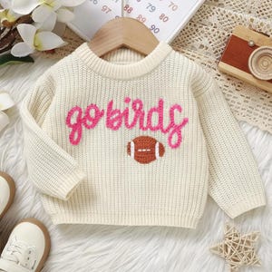 May include: Cream-colored knit sweater with the words "go birds" in pink script and a brown football graphic. The sweater has a round neckline and long sleeves. The garment is displayed on a wooden hanger.
