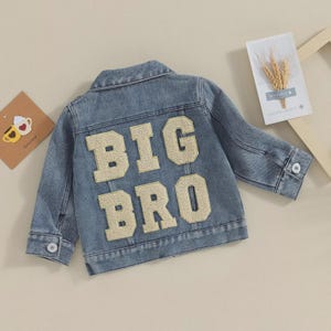 May include: Light blue denim jacket with the words "BIG BRO" in large, gold-colored letters on the back. The jacket has a classic collar, button closures, and pockets. A stylish piece for a child.
