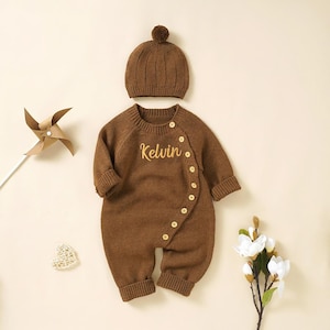 Puede incluir: Conjunto de bebé de punto marrón que incluye un pelele y un gorro a juego. El pelele tiene cierre de botones y el nombre "Kelvin" bordado en dorado. El gorro tiene un diseño acanalado y un pequeño pompón. Un conjunto acogedor para bebé.