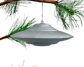 May include: A silver UFO-shaped Christmas ornament hanging from a pine tree branch.