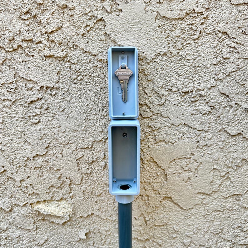 May include: A gray plastic key holder with a key inside. The key holder is mounted on a gray pole.