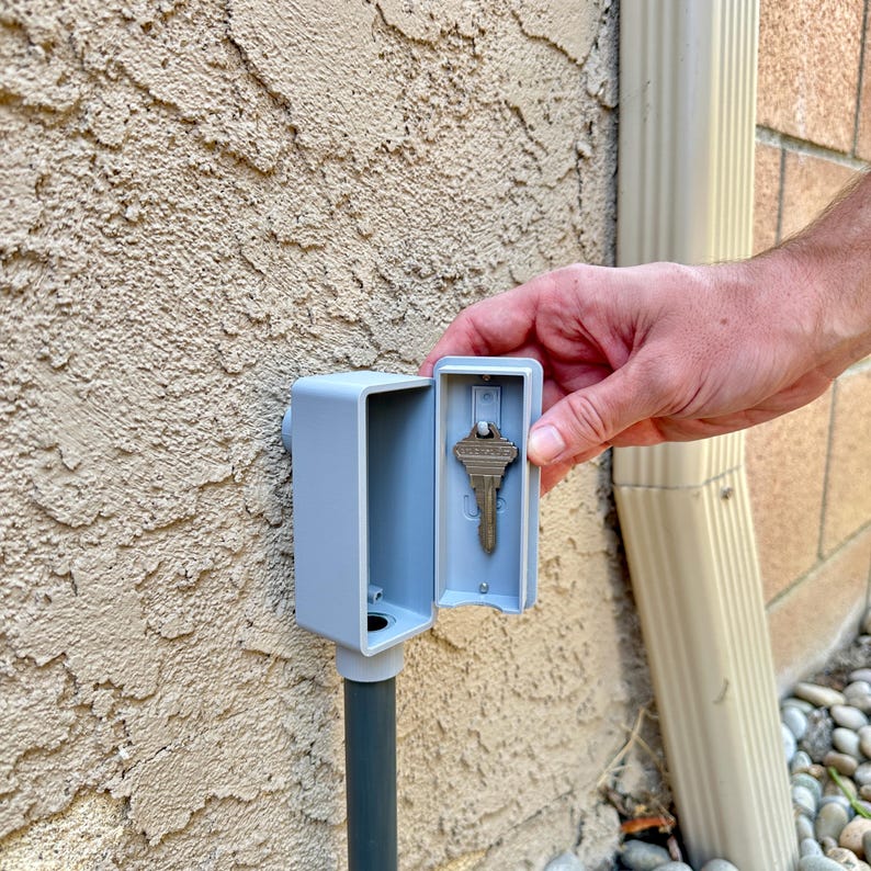 May include: A gray plastic key holder mounted to a wall with a key inside. The holder has a hinged lid that is open.