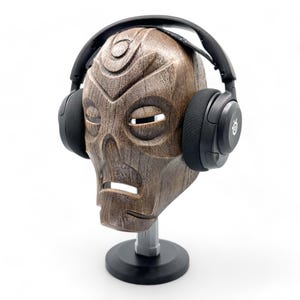 May include: A wooden mask with a carved face, mounted on a stand, and topped with black headphones. The mask has a weathered, brown finish and is set against a white background. The headphones have black earcups and a black headband.