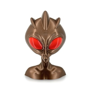 May include: A bronze-coloured alien bust with red eyes. The alien has a pointed head and a long, thin neck.