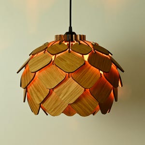 Könnte beinhalten: Eine Holz-Pendelleuchte mit einem einzigartigen Design, das an eine Kiefernzapfen erinnert. Die Leuchte besteht aus vielen einzelnen Holzstücken, die in einem kreisförmigen Muster angeordnet sind. Die Leuchte wird mit einem warmen, orangenen Licht beleuchtet.