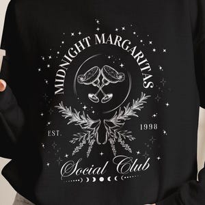 May include: A black long-sleeve sweatshirt featuring a white graphic print that reads Midnight Margaritas Social Club, Est. 1998, with illustrations of two clinking margarita glasses, leafy branches, stars, and a moon phase cycle.