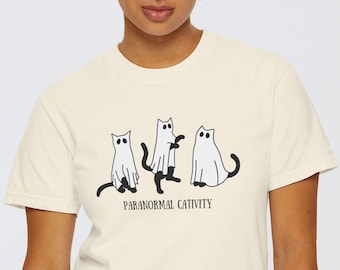 May include: A cream colored t-shirt with a graphic of three white cats with black eyes and tails, appearing as ghosts. The text "Paranormal Cat-tivity" is printed below the graphic.