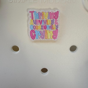 K&ouml;nnte beinhalten: Eine wei&szlig;e Tragetasche mit dem gepr&auml;gten Text "SIMPLE MODERN" oben. Auf der Tasche ist ein farbenfroher, glitzernder Aufkleber angebracht, der die Worte "Thriving, Surviving & Occasionally Crying" in einer verspielten Schriftart zeigt. Die Tasche hat mehrere kreisf&ouml;rmige L&ouml;cher.
