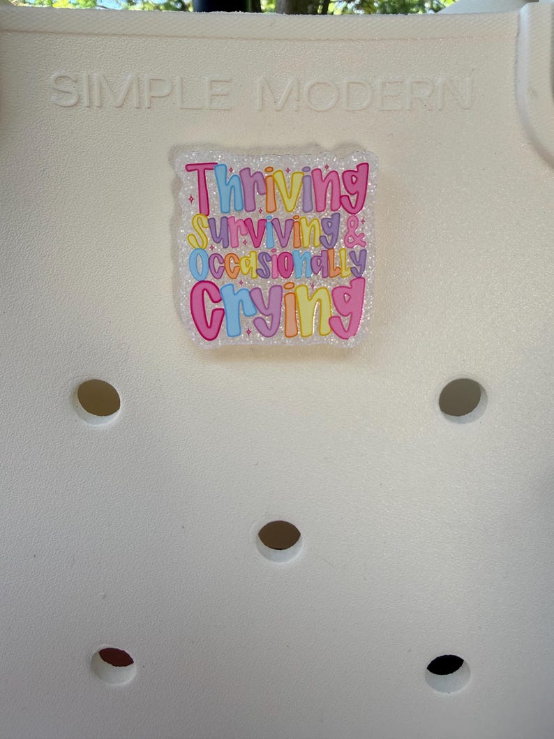 K&ouml;nnte beinhalten: Eine wei&szlig;e Tragetasche mit dem gepr&auml;gten Text "SIMPLE MODERN" oben. Auf der Tasche ist ein farbenfroher, glitzernder Aufkleber angebracht, der die Worte "Thriving, Surviving & Occasionally Crying" in einer verspielten Schriftart zeigt. Die Tasche hat mehrere kreisf&ouml;rmige L&ouml;cher.